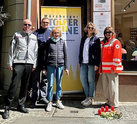 Fünf Personen vor einem Rollup mit der Aufschrift "Stolpersteine". zu ihren Füßen vier kerzen und Blumen rund um den neu verlegten Stolperstein.